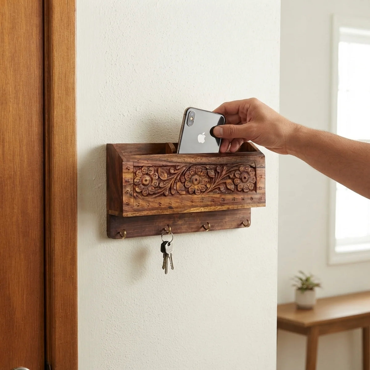 Hand carved wooden wall organizer beside a door, holding a phone in the top compartment with house keys on the hooks